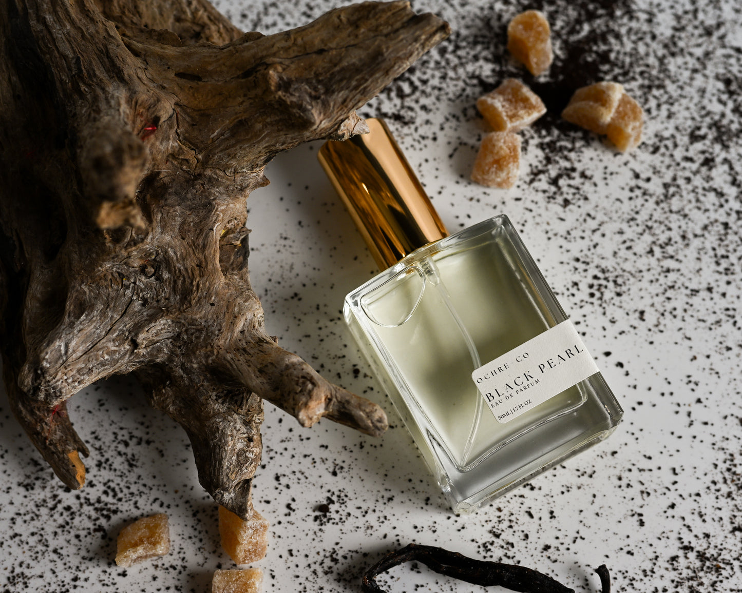 Perfume bottle of Black Pearl on a white reflective surface, driftwood, vanilla bean, and white background