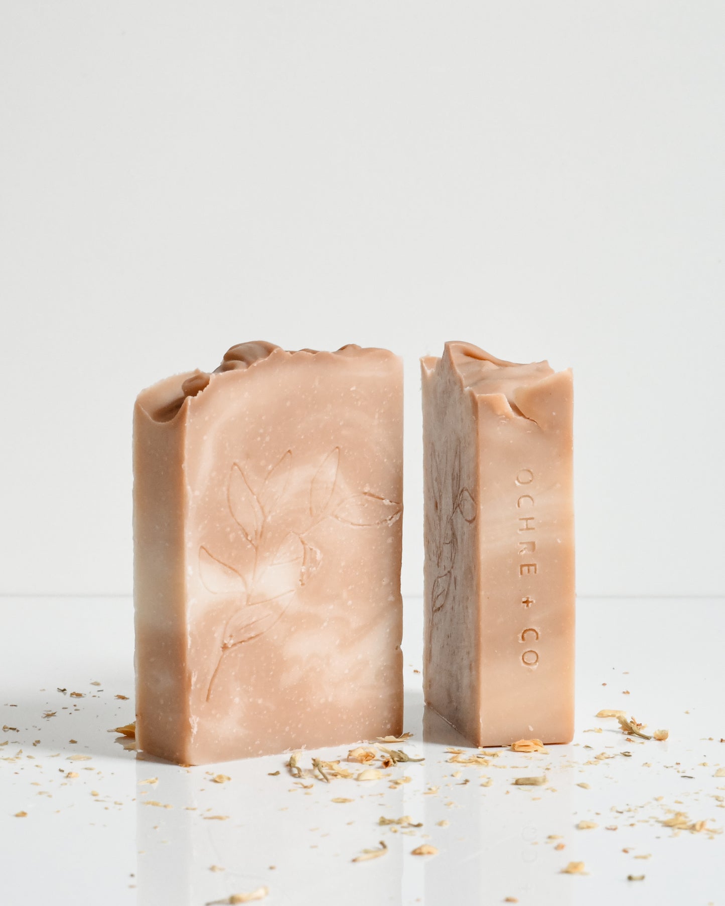 Two bar soaps with a beige color and a textured appearance, with the brand 'Ochre + Co' visible on one of the soaps.