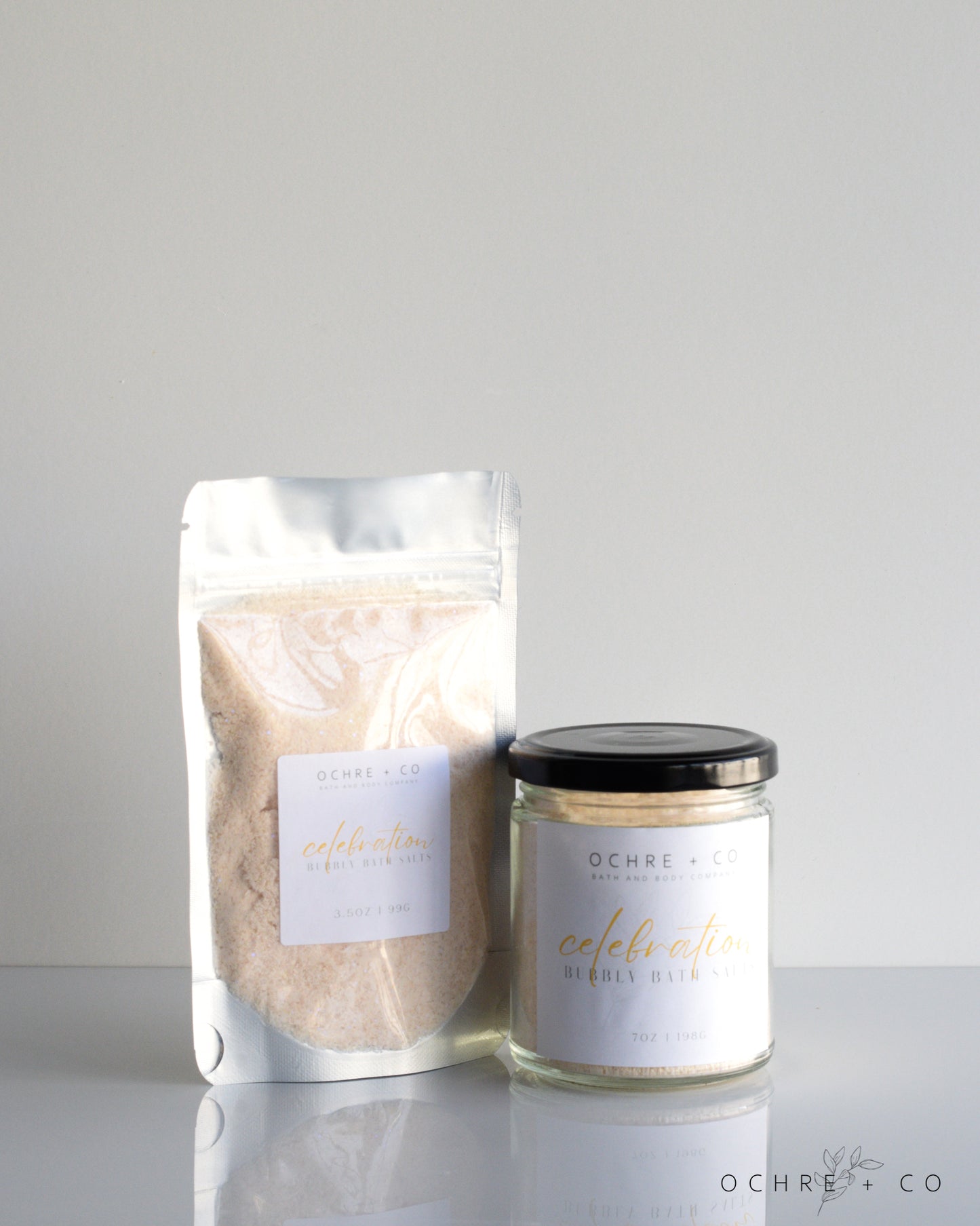 A jar and a transparent package containing white bath salts, with labels in both items. The jar is smaller and placed in front of the package.