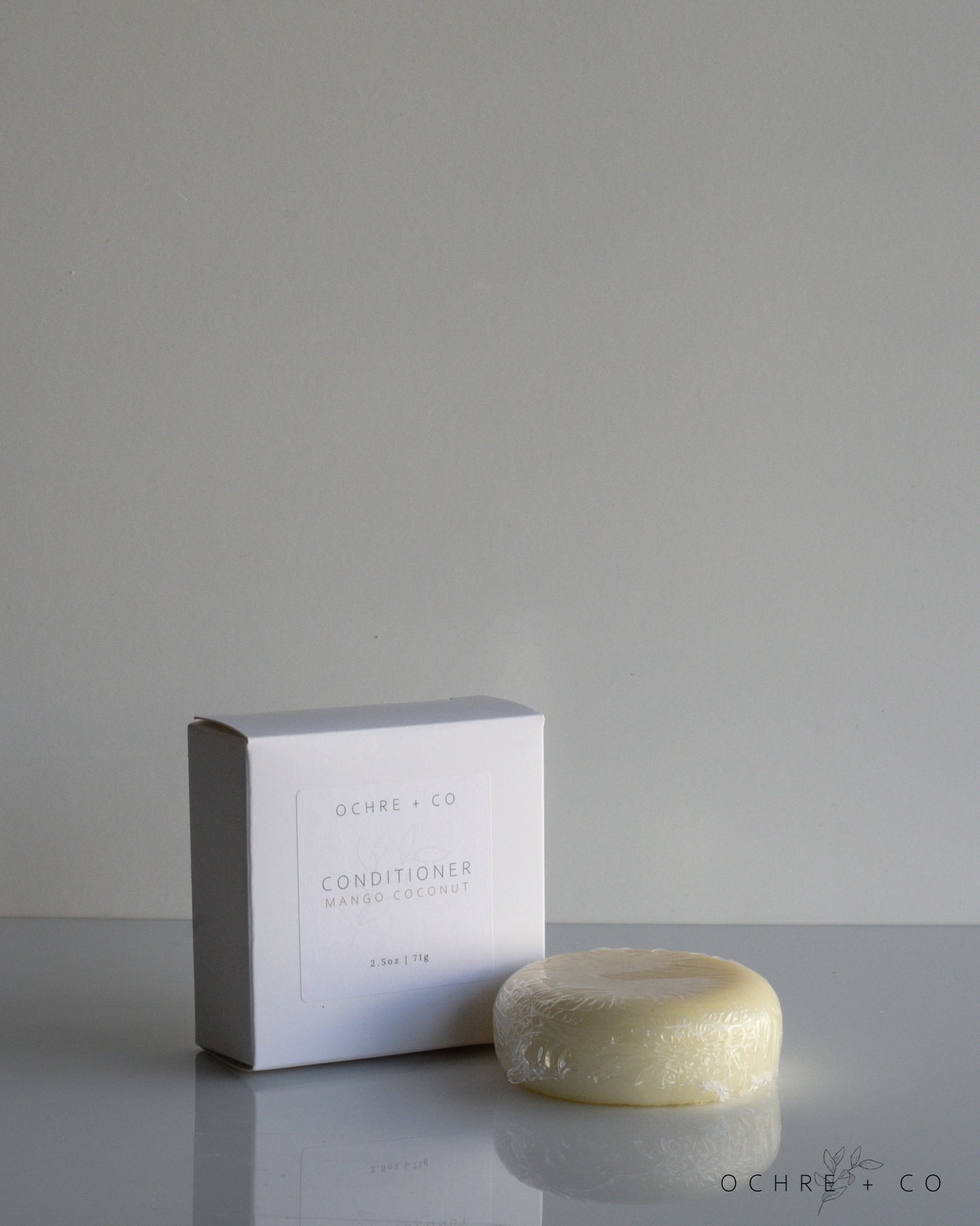 A conditioner bar next to its packaging box, labeled Mango Coconut Conditioner. The box is white with text, and the bar appears beige.