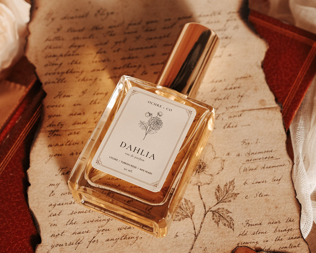 A bottle of Dahlia perfume by Ochre & Co. resting on an aged, handwritten paper sheet over a vintage leather journal, surrounded by soft white fabric and a rose.