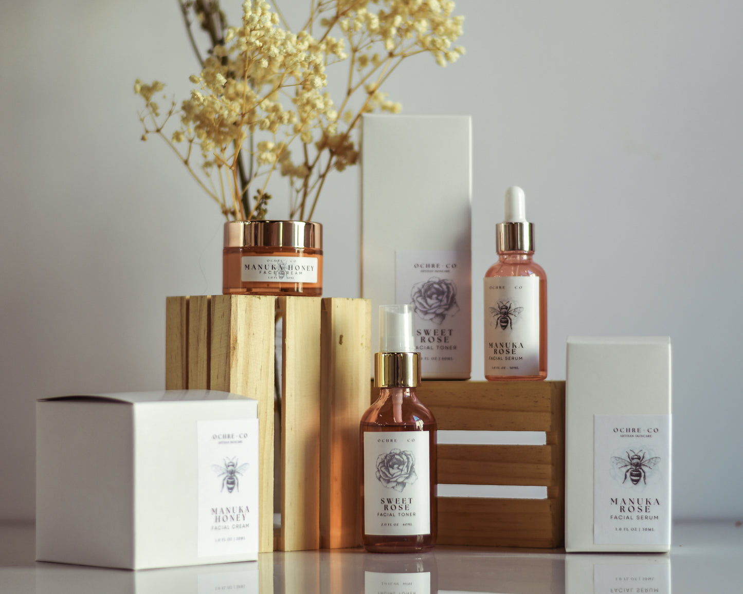 A group photo of Sweet Rose toner, Manuka Honey facial cream, and Manuka Rose facial serum on a white background with their boxes, wooden crates for height, and dried baby's breath florals