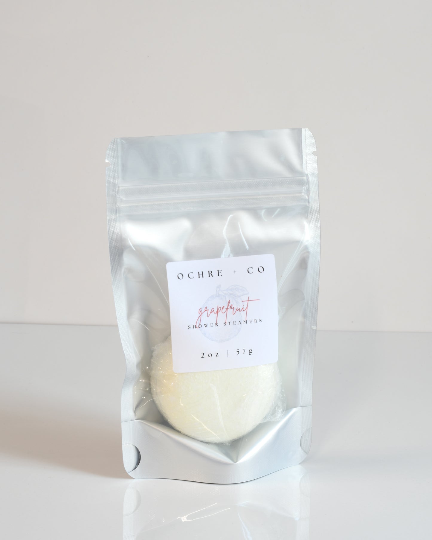 A shower steamer packaged in a clear plastic bag with a label reading 'Grapefruit Shower Steamer', with a white, solid-colored steamer visible inside.