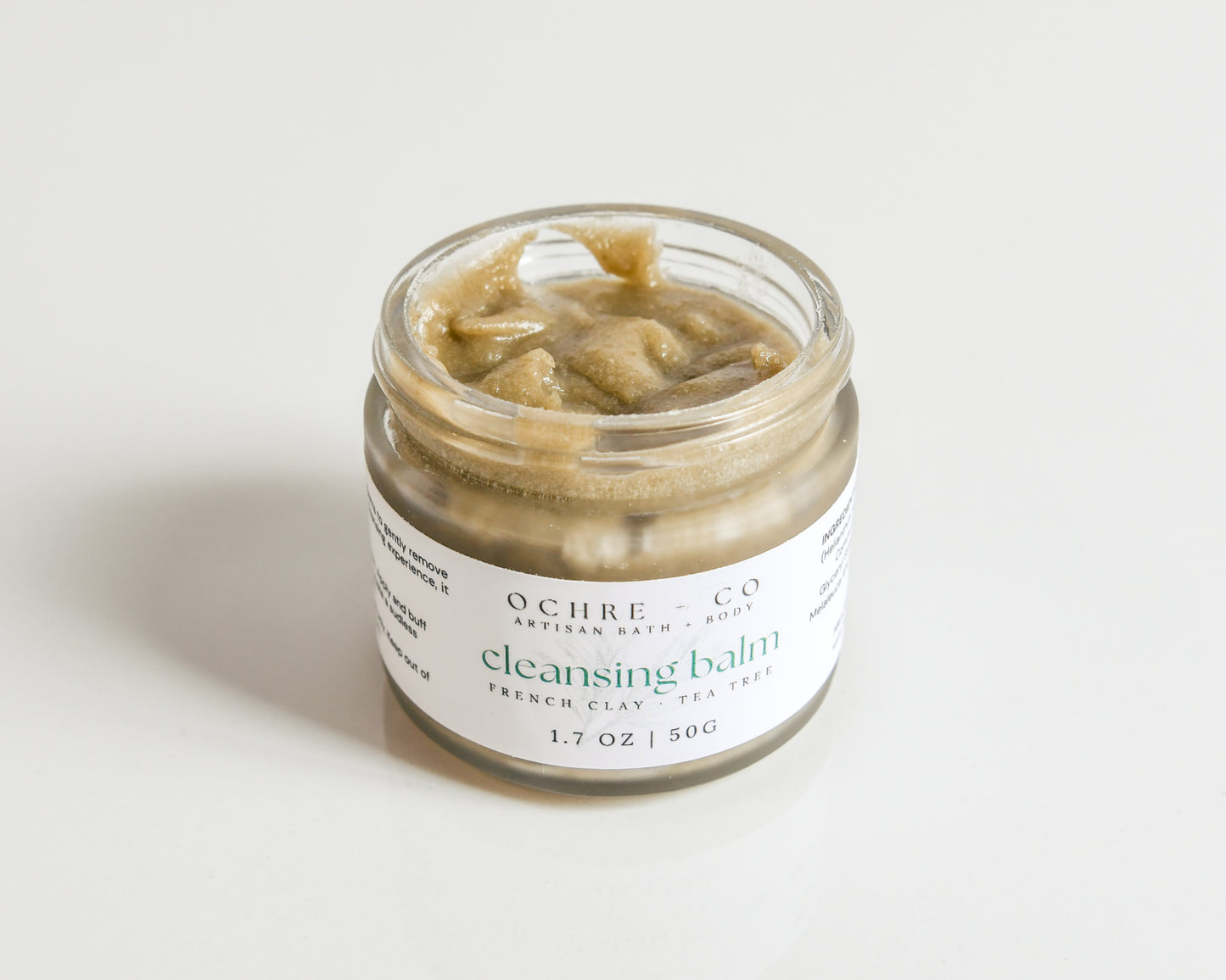A small jar of Facial Cleansing Balm with tea tree essential oil and French green clay without a lid showcasing the creamy texture on a white background.