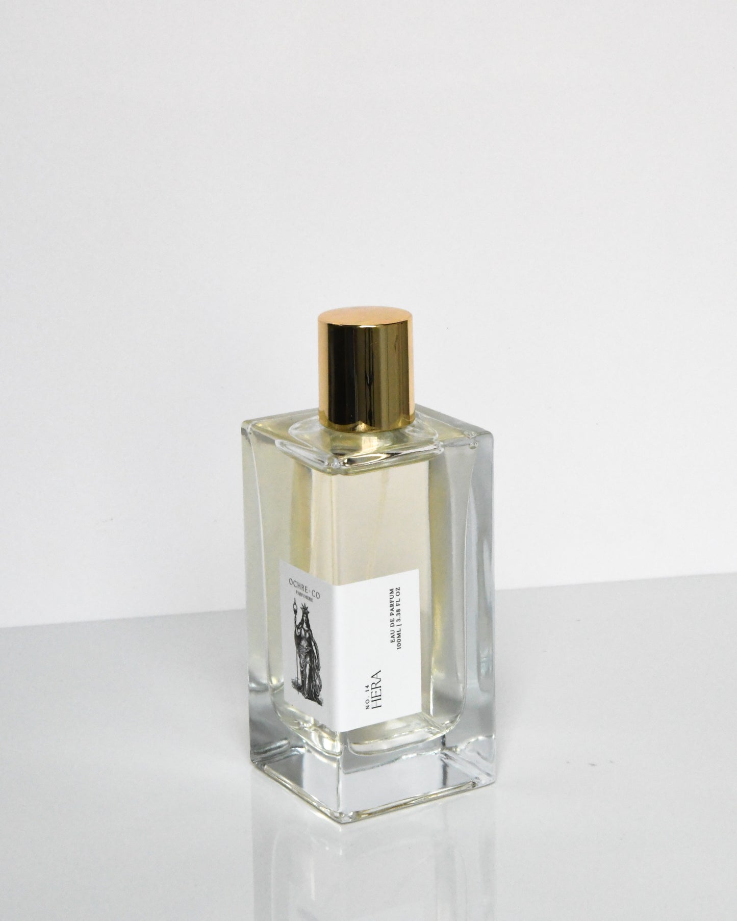 A clear glass bottle of Hera Eau de Parfum with a gold-colored cap. The label features an illustration of a woman.