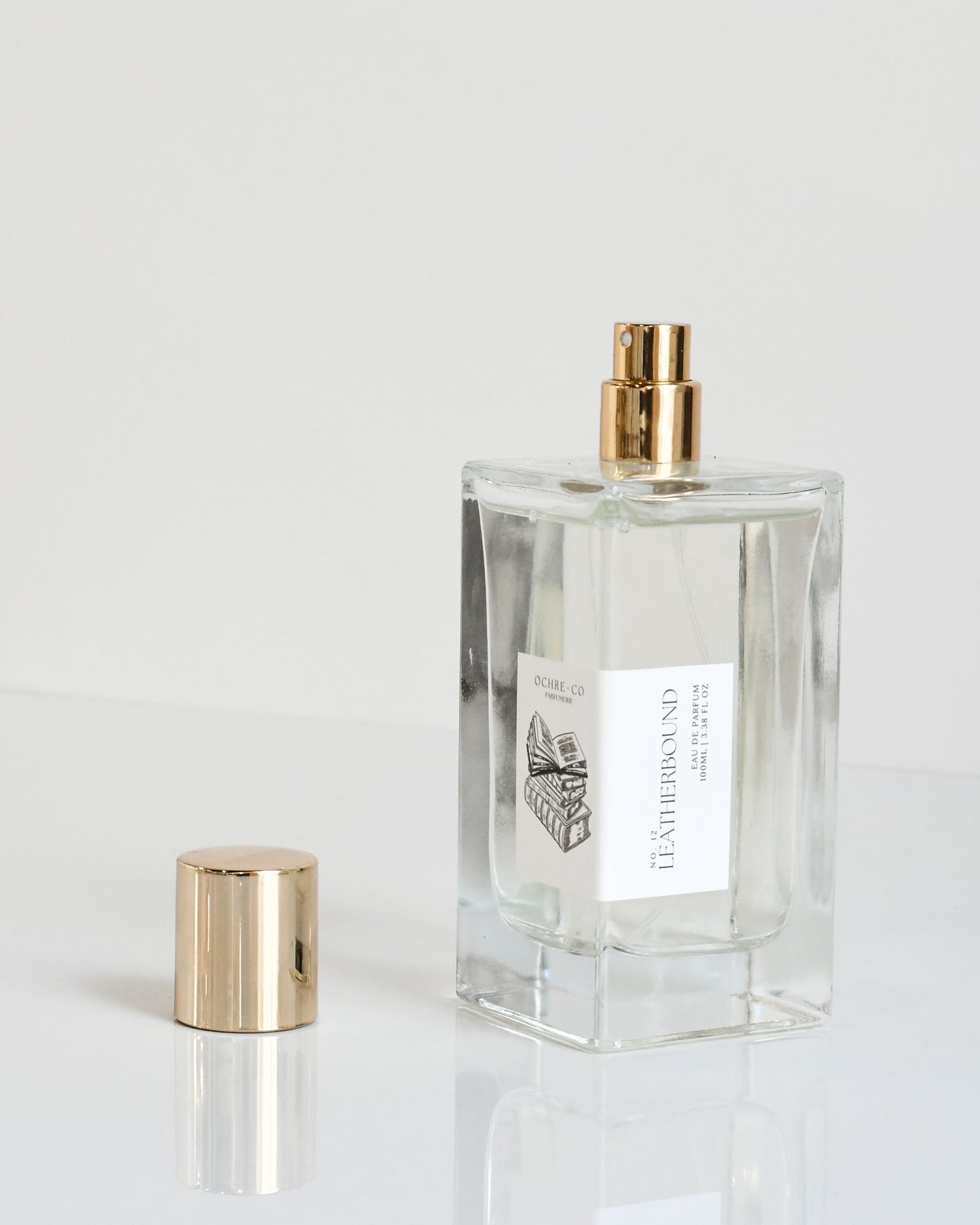 A clear glass bottle of 'Leatherbound - Eau de Parfum' with a gold-colored spray nozzle and cap removed, placed next to its matching gold cap on a white surface.