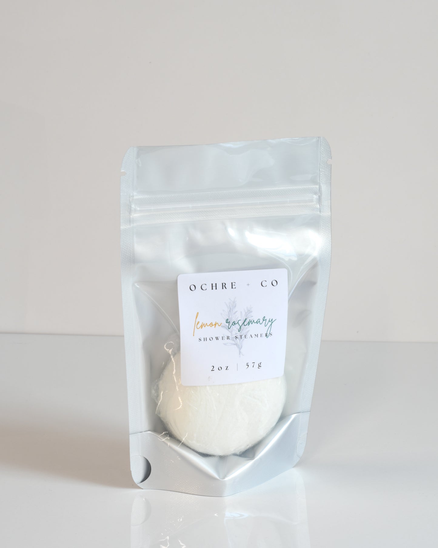 A shower steamer packaged in a clear plastic pouch with a label reading 'Lemon Rosemary' from the brand OCHRE + CO.
