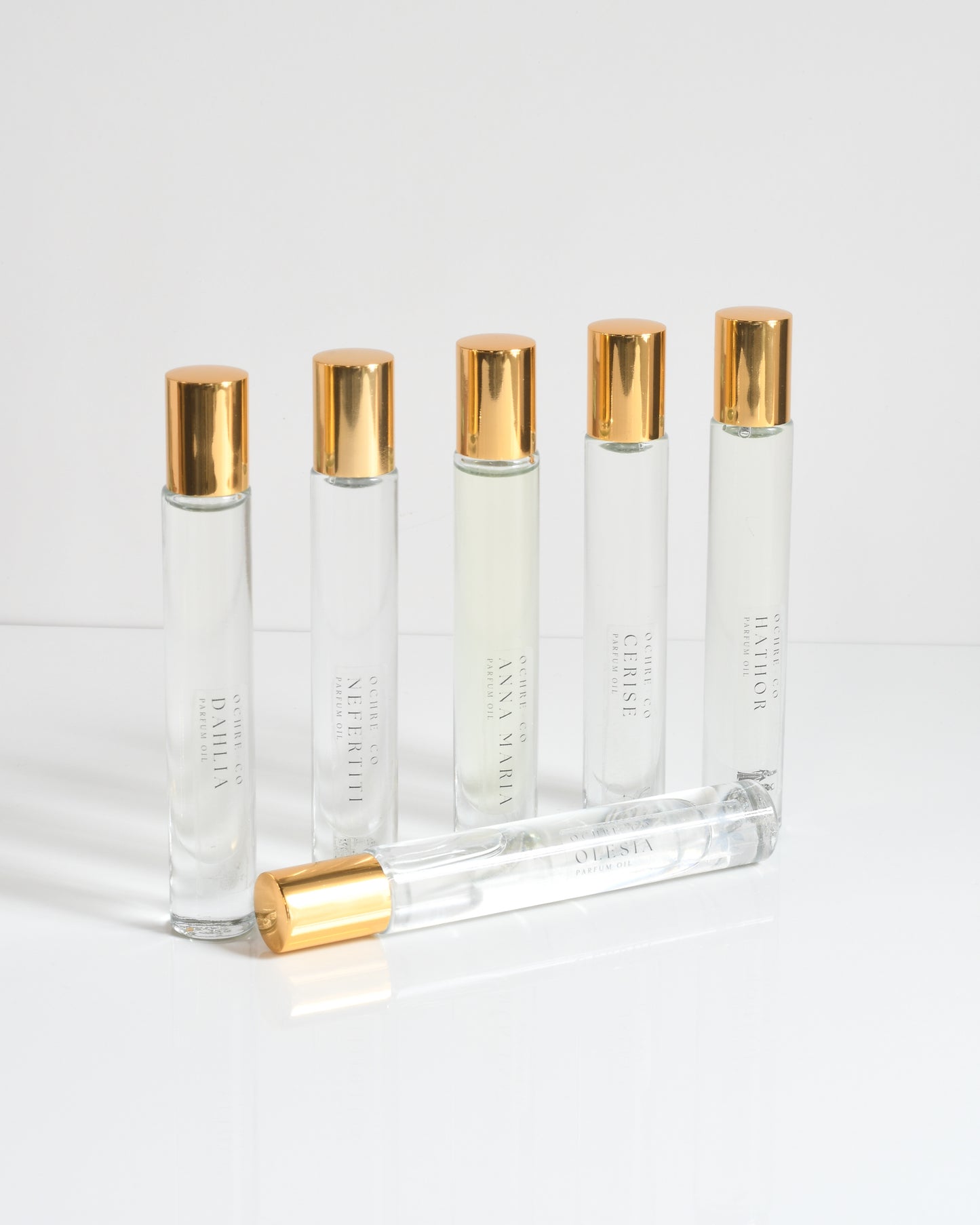 A grouping of six perfume oils in clear roller ball containers with gold lids on a white background