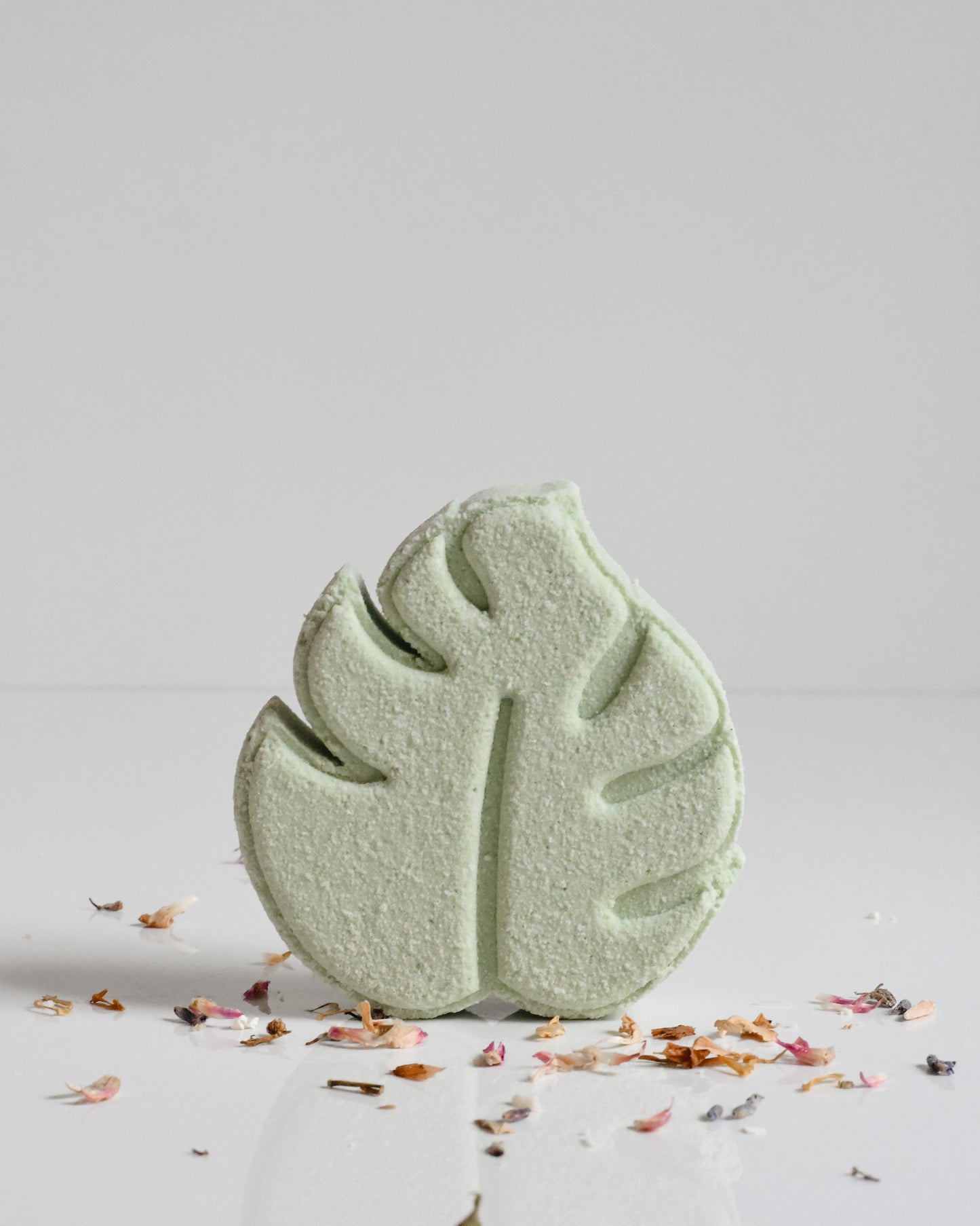 A green bath bomb with a leaf pattern on top, surrounded by scattered flower petals on a white surface.