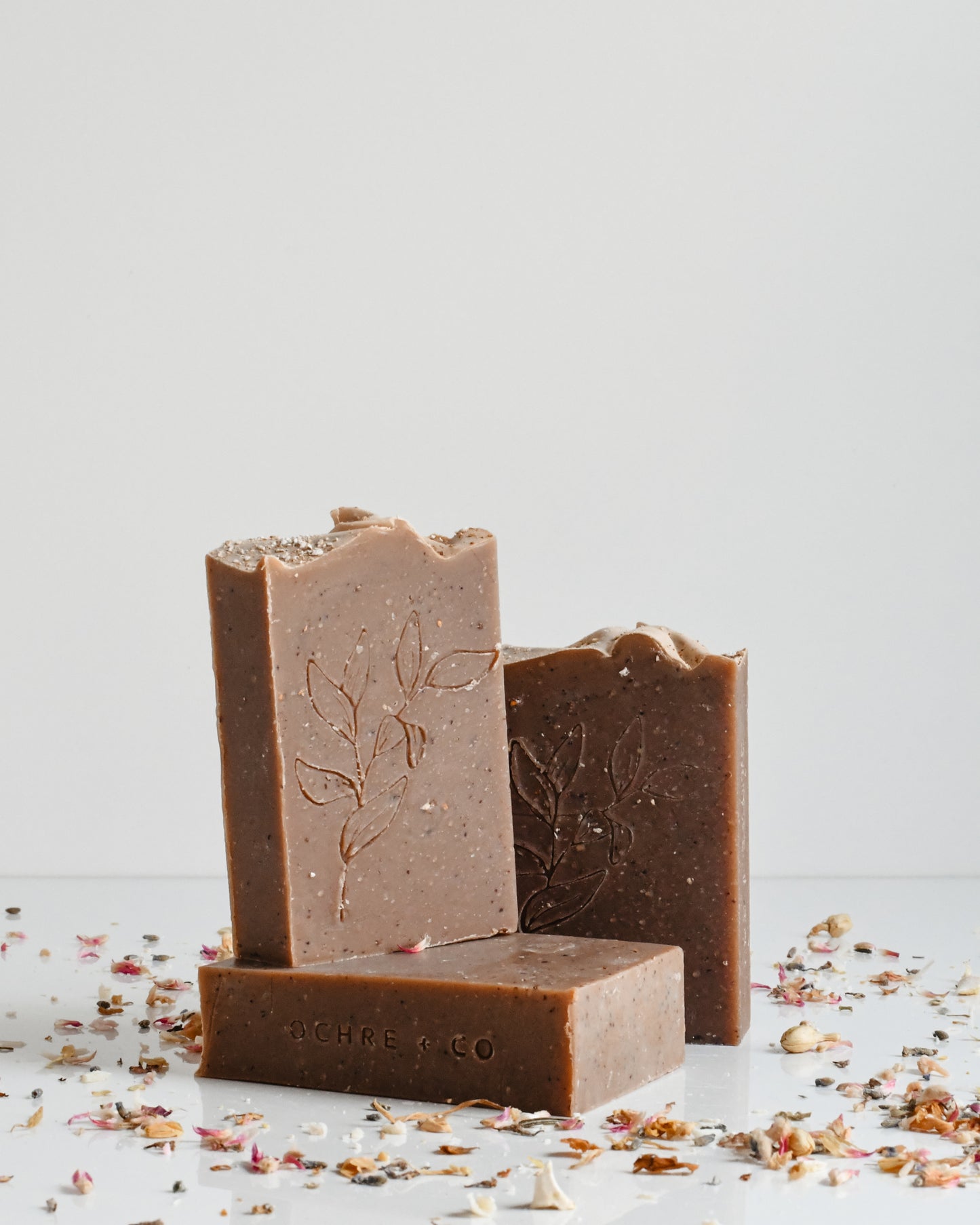 three arranged bars of speckled brown soap surrounded by scattered dried florals on a white background