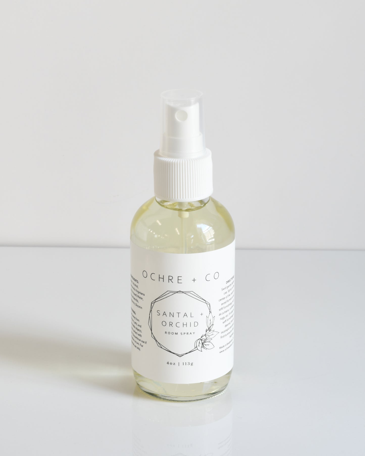 A clear glass bottle with a white spray nozzle labeled 'Santal + Orchid Room Spray' on a white background