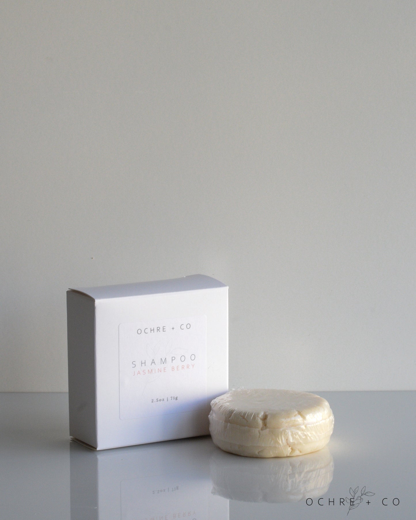 A shampoo bar next to its white packaging box, labeled 'JASMINE BERRY', on a reflective surface.