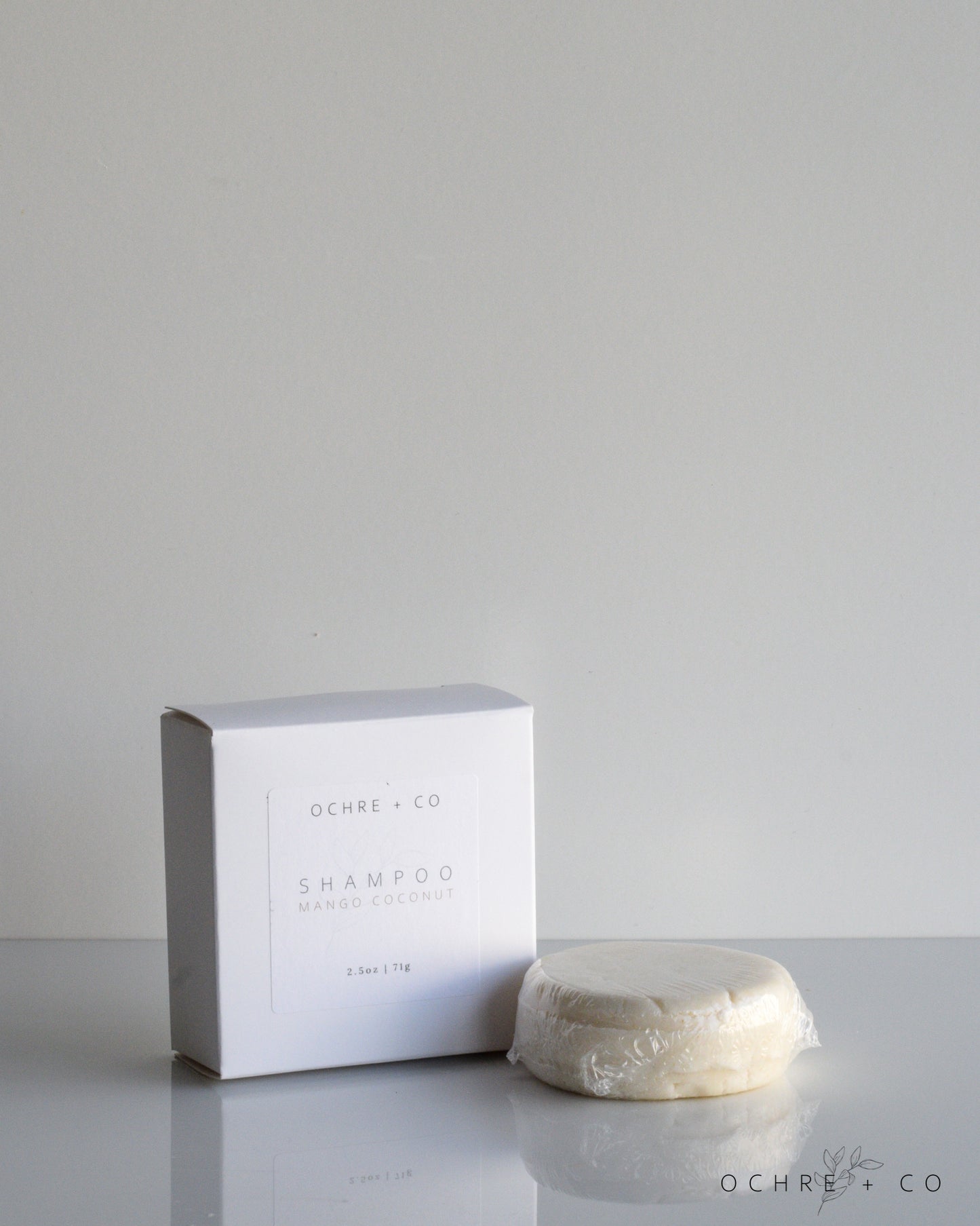A mango coconut shampoo bar in front of its white packaging box, with the brand name 'OCHRE + CO' visible on the box. The bar itself is cream-colored and appears solid, set against a grey background.