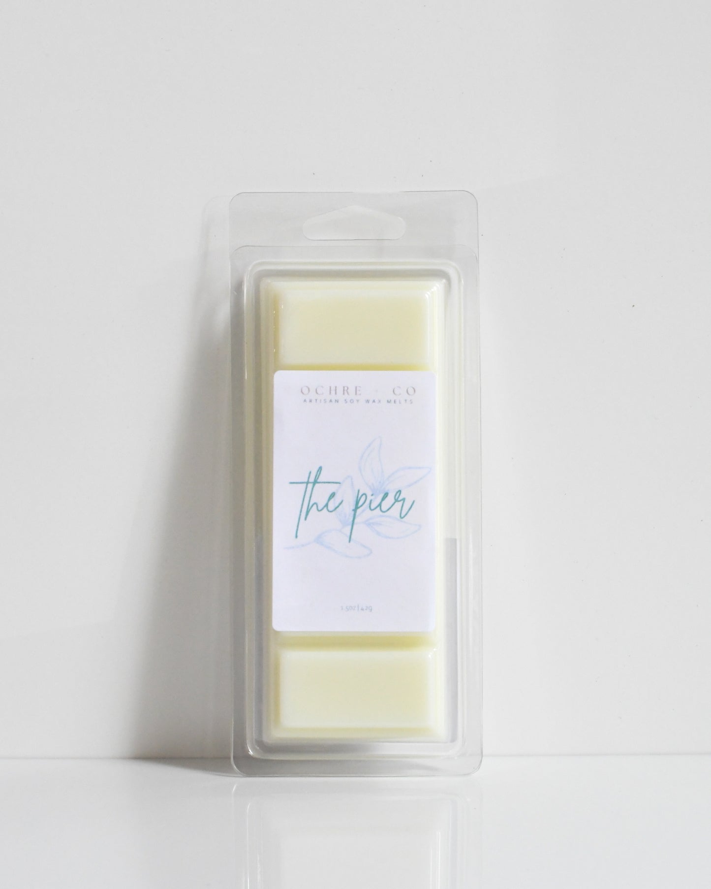 A white wax melt on a white background in its plastic packaging, the label reads "THE PIER"