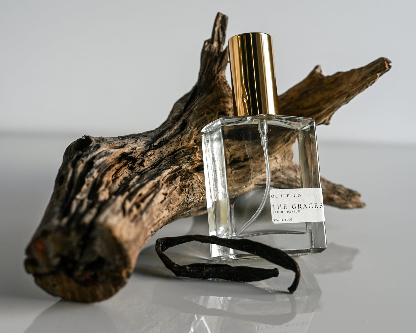 Perfume bottle of The Graces with gold cap and clear label on a wooden log