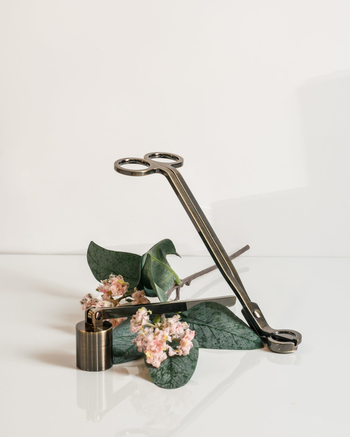 A rubbed bronze candle snuffer and wick trimmer with a piece of decorative floral on a white background