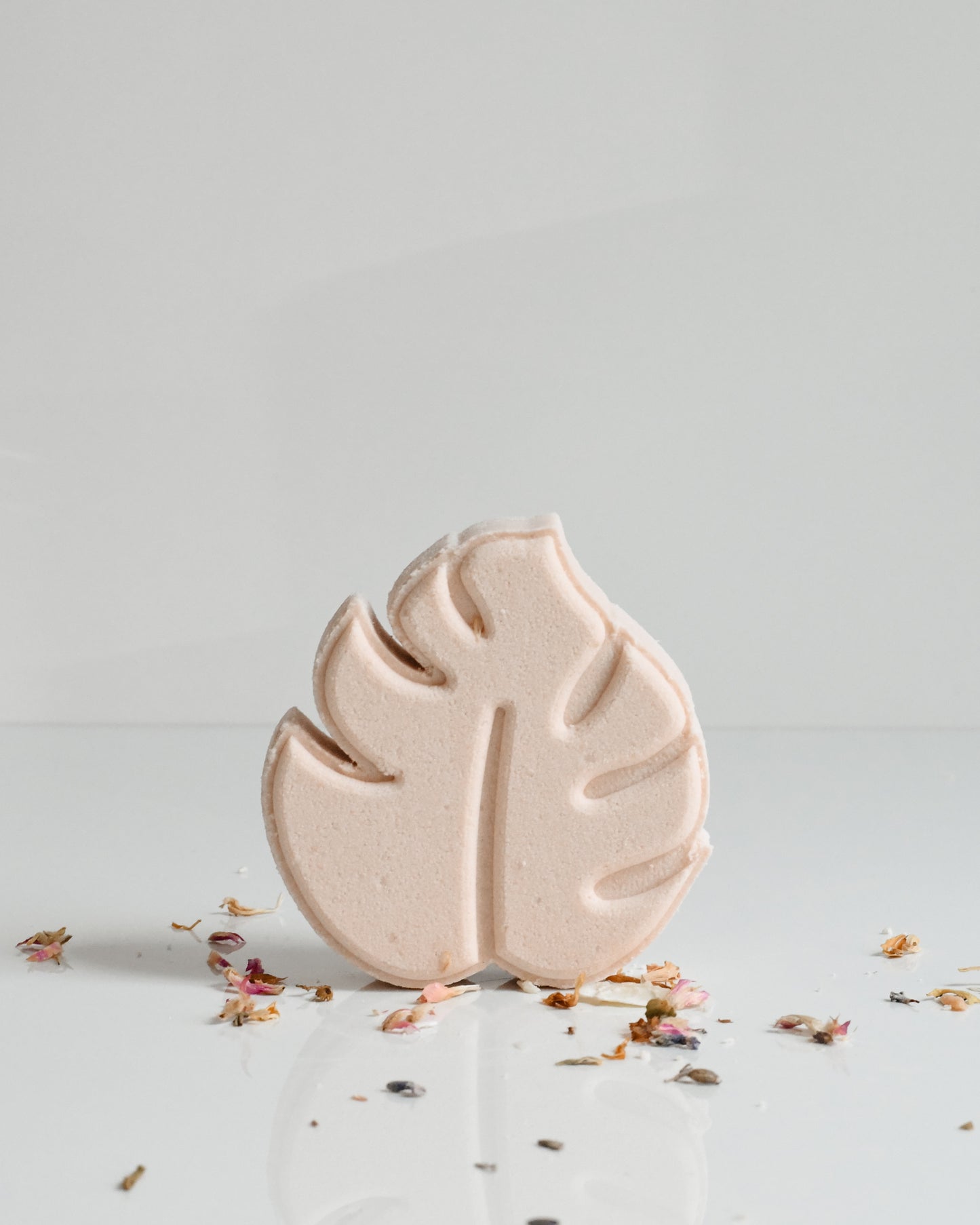 A light beige bath bomb with a leaf pattern on top, surrounded by scattered flower petals on a white surface.