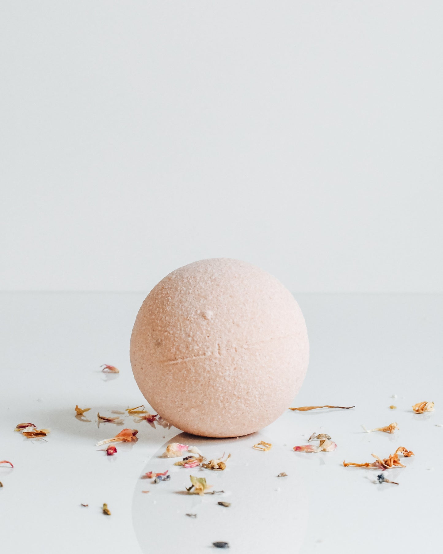 A soft pink spherical bath bomb on a white background with scattered dried florals around it