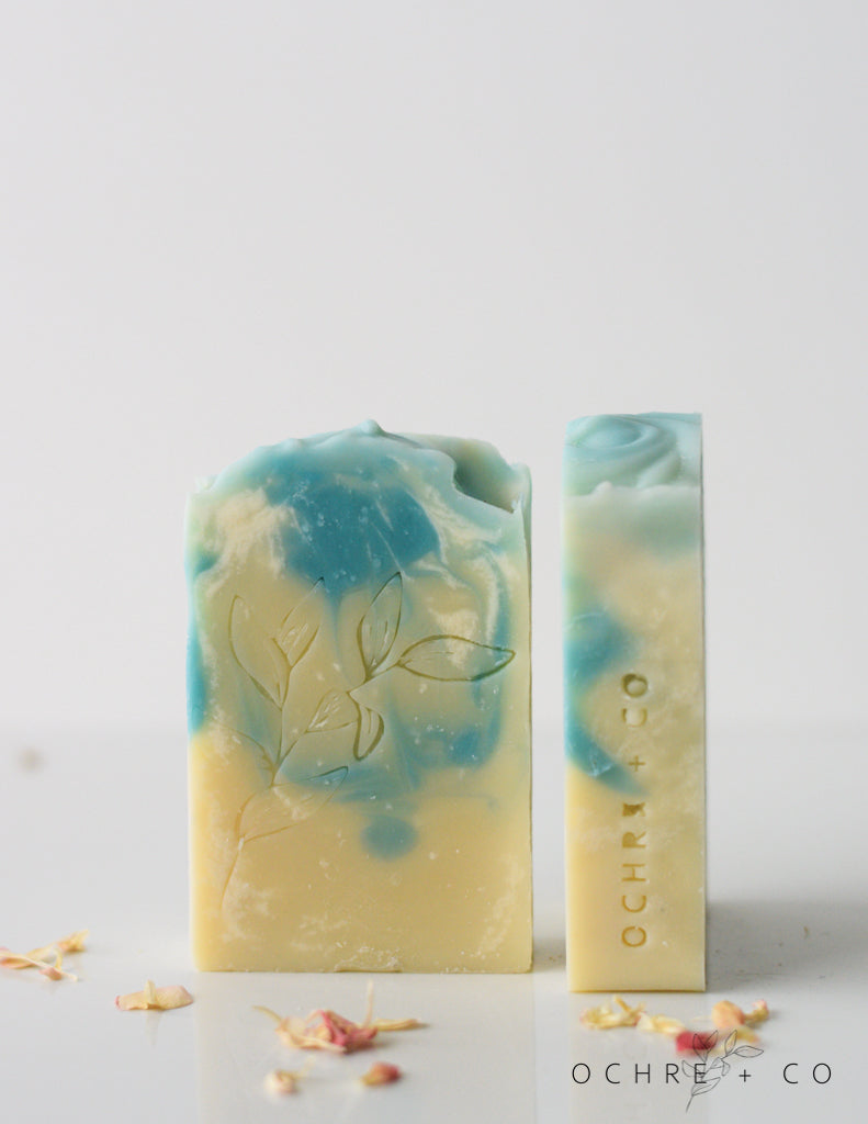 Two Ocean Mist light blue and white swirled soap bars with a swirl design on top, with a single bar leaning against the stack, and small dried flowers scattered around.