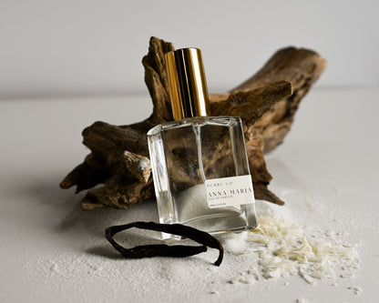 Perfume bottle of Anna Maria with gold cap and clear label on a light surface with driftwood and vanilla bean.