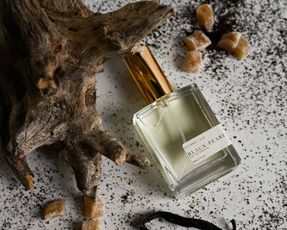 Perfume bottle of Black Pearl on a white reflective surface, driftwood, vanilla bean, and white background