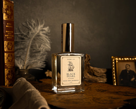 A bottle of Black Pearl eau de partfum stands on a rustic wooden surface, surrounded by vintage props including an antique book, a feather quill, a compass, and a framed portrait in a dimly lit setting.