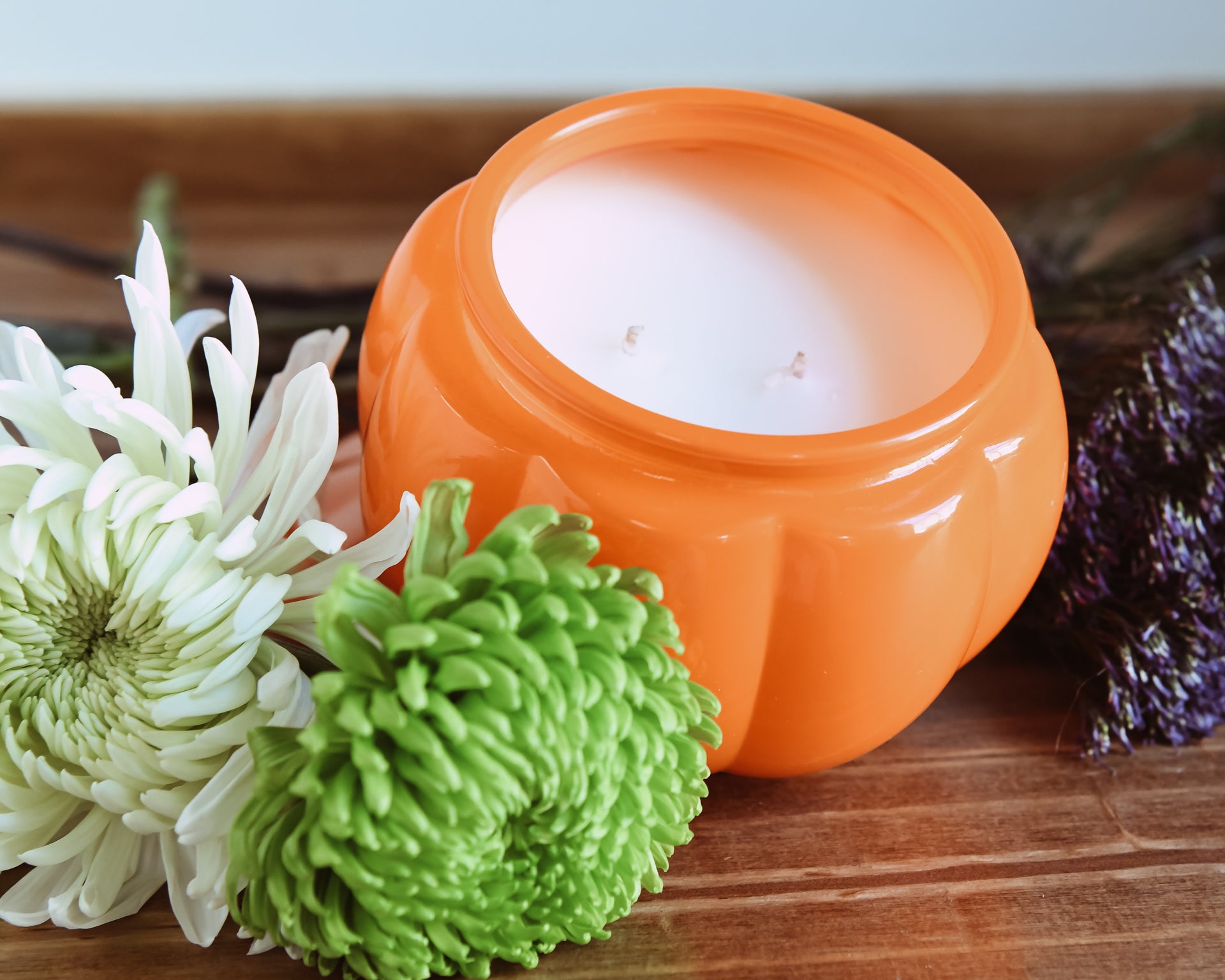 An orange pumpkin-shaped ceramic candle vessel on a wooden counter with white and green mums and purple florals