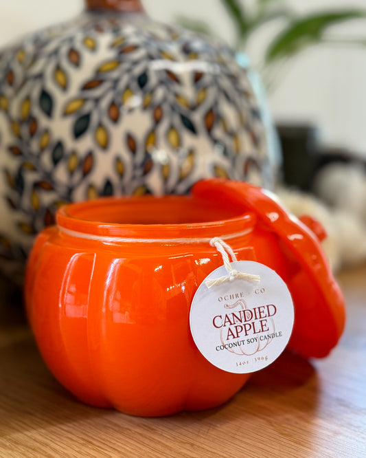 An orange pumpkin-shaped coconut soy candle with a label that reads 'Candied Apple'. The candle is placed on a wooden surface and has a decorative item in the background.