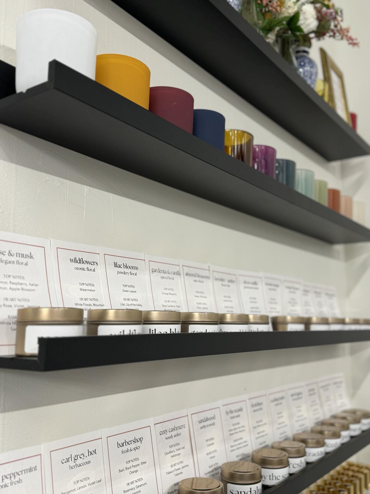 A wall with black shelves holding candle class materials including various colored candle vessels, tins with scented wax, and notes for the scents