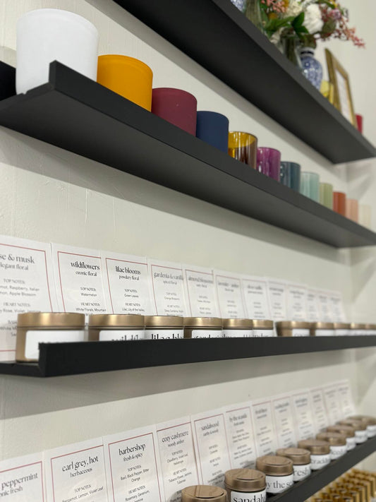 A wall with black shelves holding candle class materials including various colored candle vessels, tins with scented wax, and notes for the scents