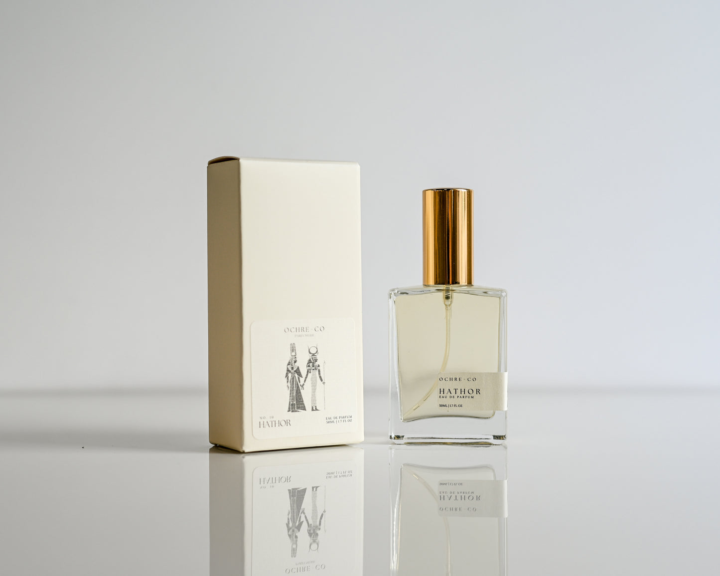 Perfume bottle of Hathor with gold cap and matching box on a reflective surface with a light gray background