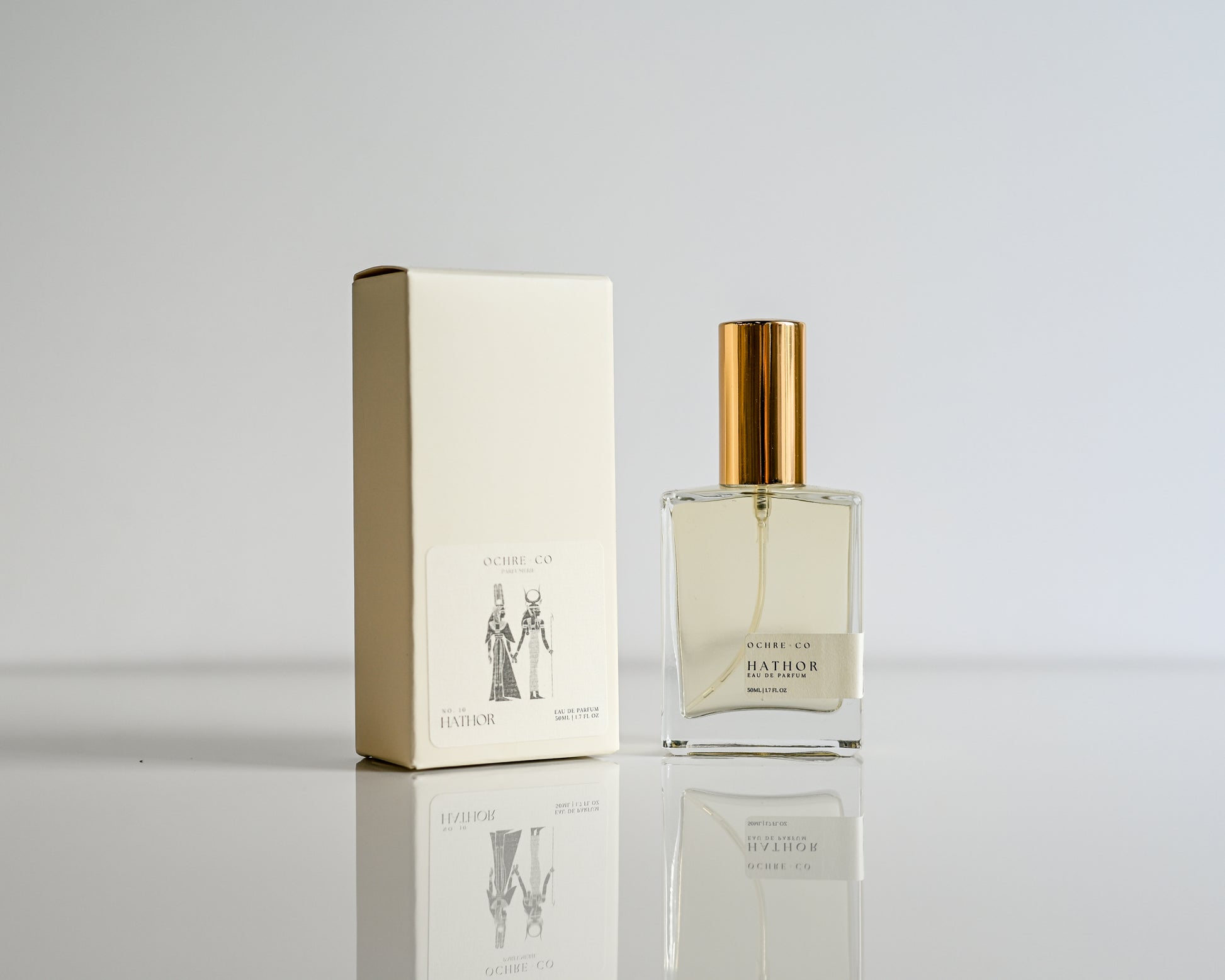 Perfume bottle of Hathor with gold cap and matching box on a reflective surface with a light gray background