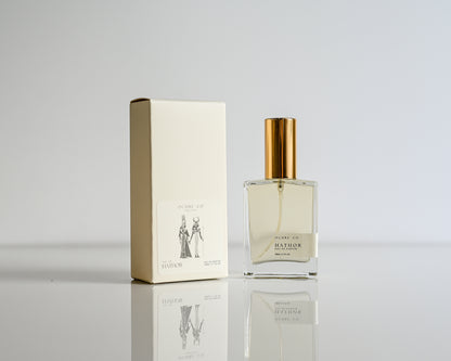 Perfume bottle of Hathor with gold cap and matching box on a reflective surface with a light gray background