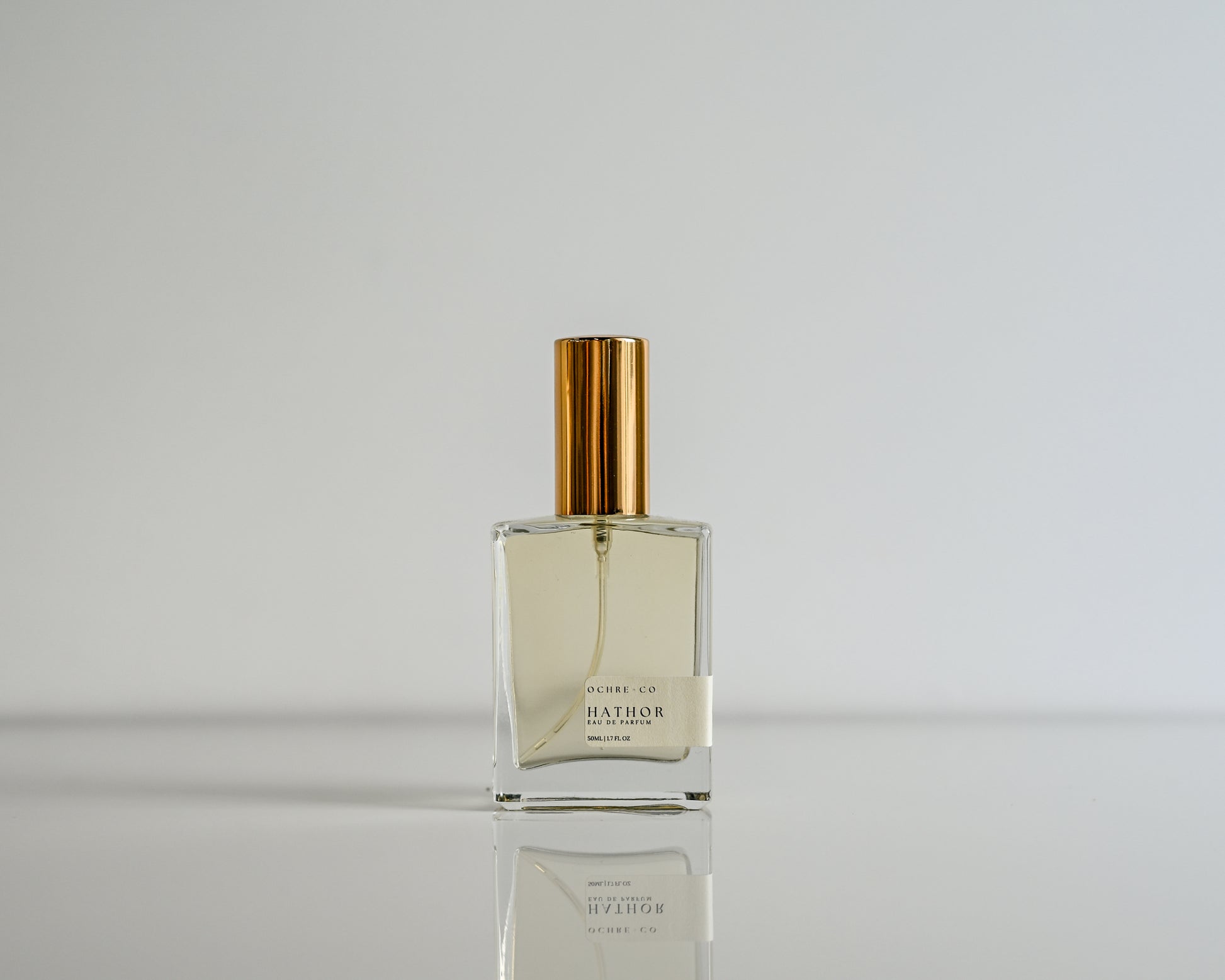 Perfume bottle of Hathor with gold cap on a reflective surface against a light gray background