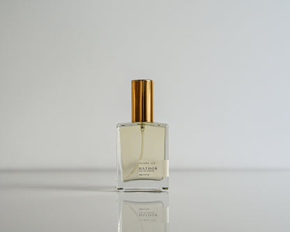 Perfume bottle of Hathor with gold cap on a reflective surface against a light gray background