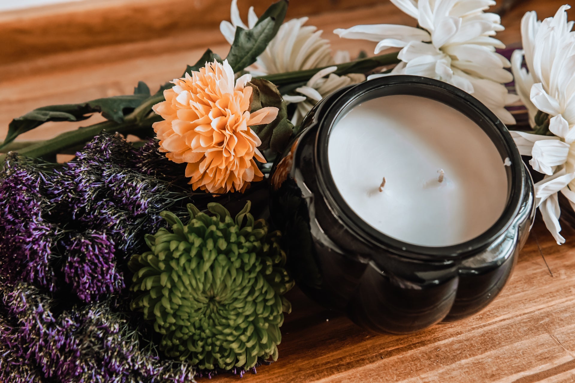 A black pumpkin-shaped ceramic candle vessel with two cotton wicks on a wooden countertop, fall-colored florals, and soft white wax