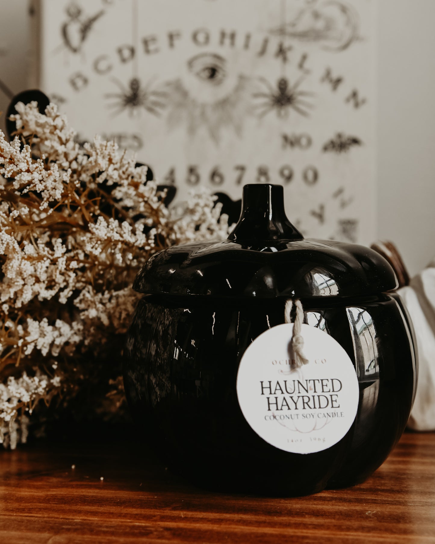 A black coconut soy candle with a label that reads 'Haunted Hayride', placed inside a black pumpkin-shaped container. The container is on a wooden surface with a decorative white item and a wall with a projector displaying letters and patterns in the background.
