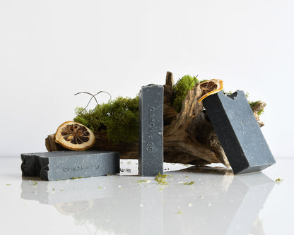 Three bars of black soap on a white surface with natural elements like moss and dried lemon slices.