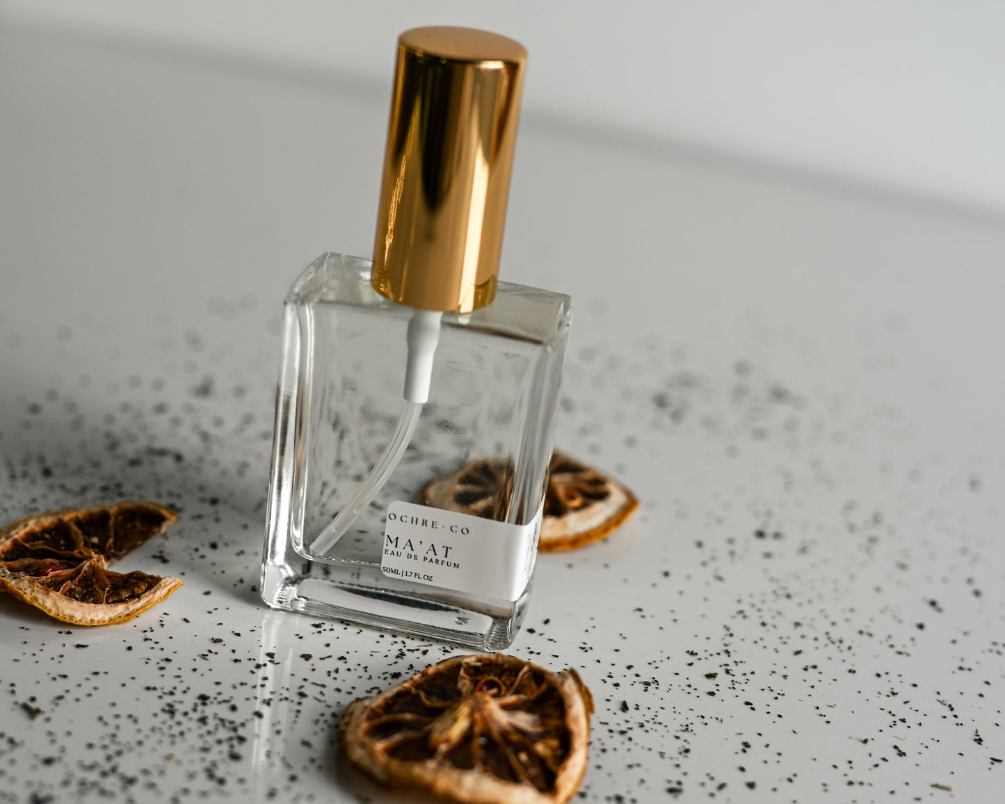 Clear perfume bottle of Ma'at with gold cap on a textured surface with dried oranges