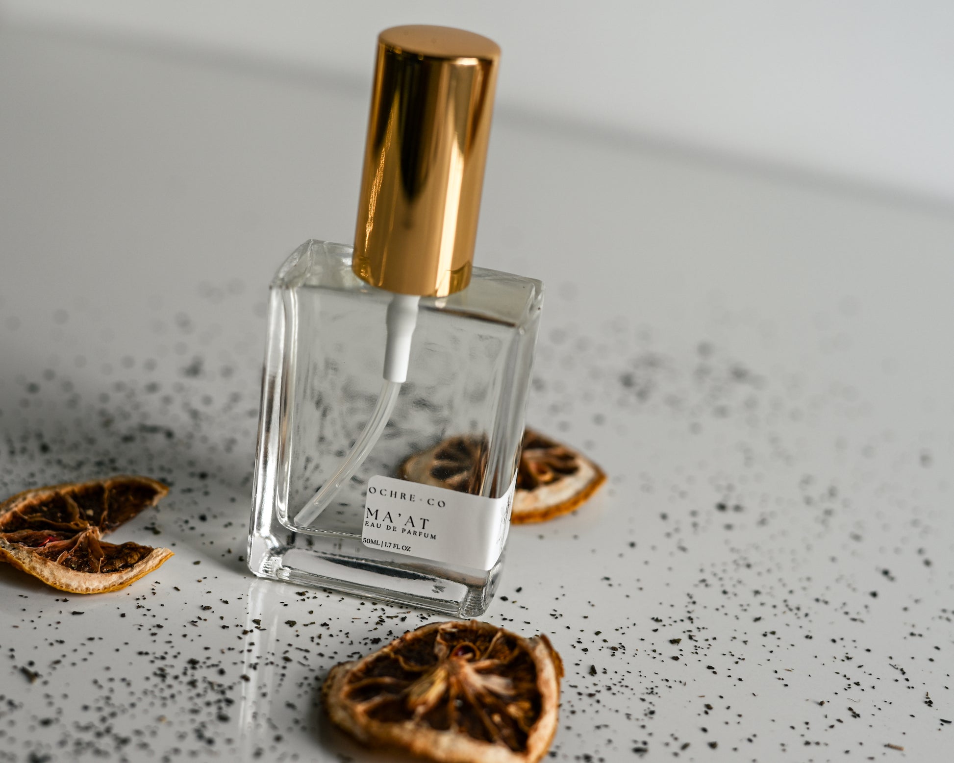 Clear perfume bottle of Ma'at with gold cap on a textured surface with dried oranges