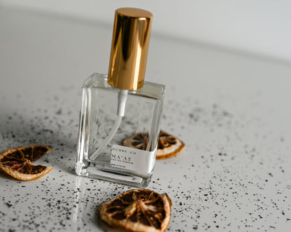 Clear perfume bottle of Ma'at with gold cap on a textured surface with dried oranges