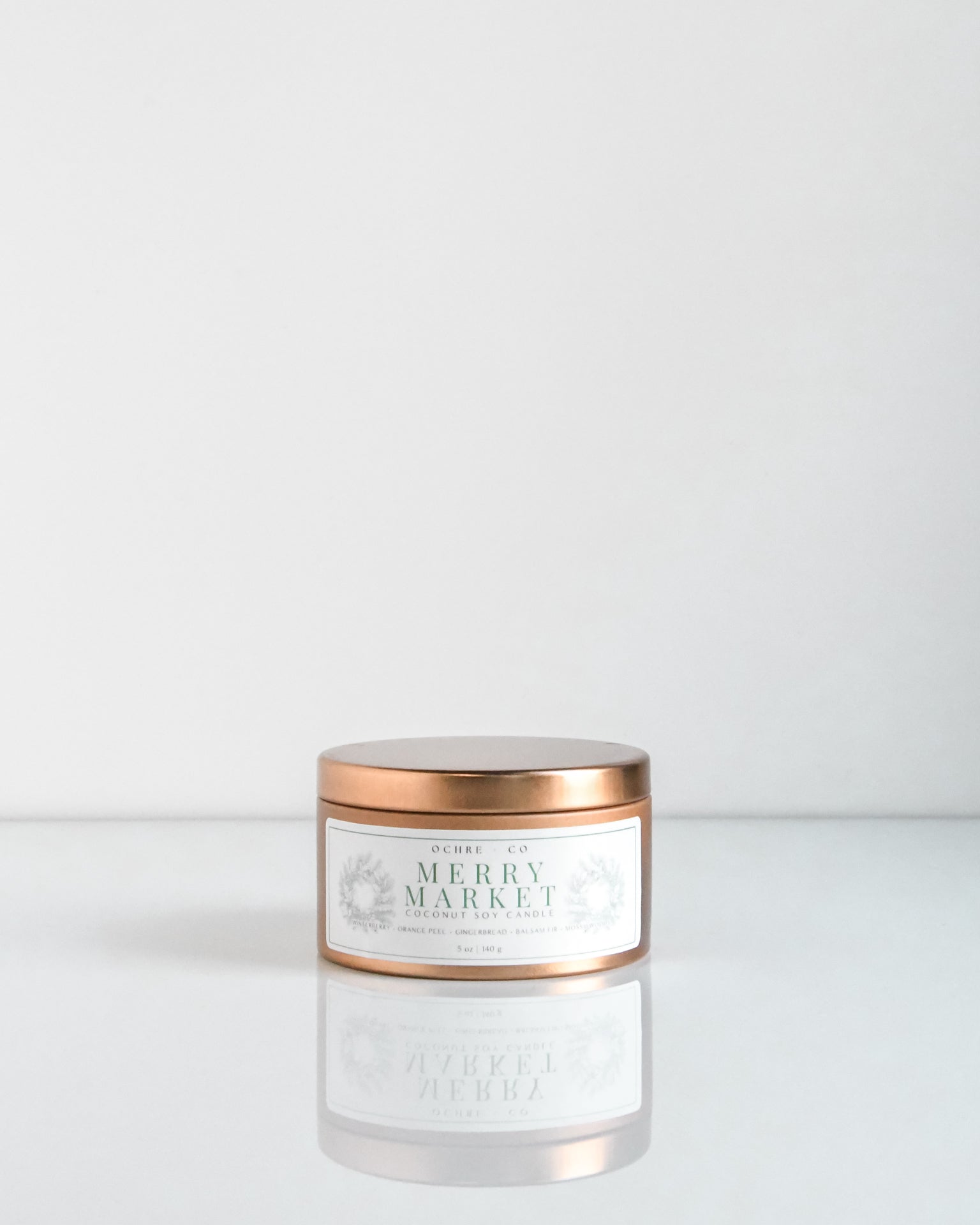 A copper metal candle vessel with the label 'Merry Market' on a white reflective surface against a white background.