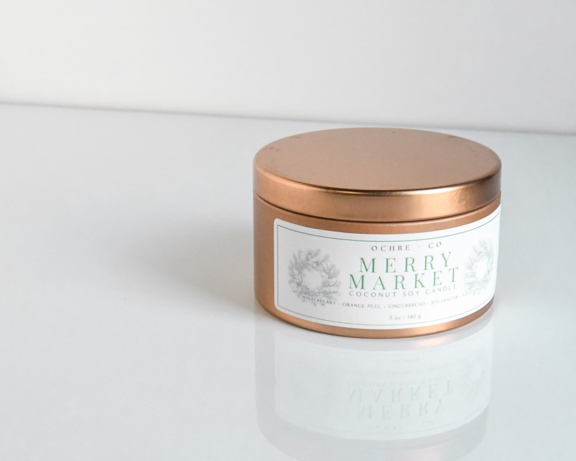 A copper metal candle vessel with the label 'Merry Market' on a white reflective surface against a white background.
