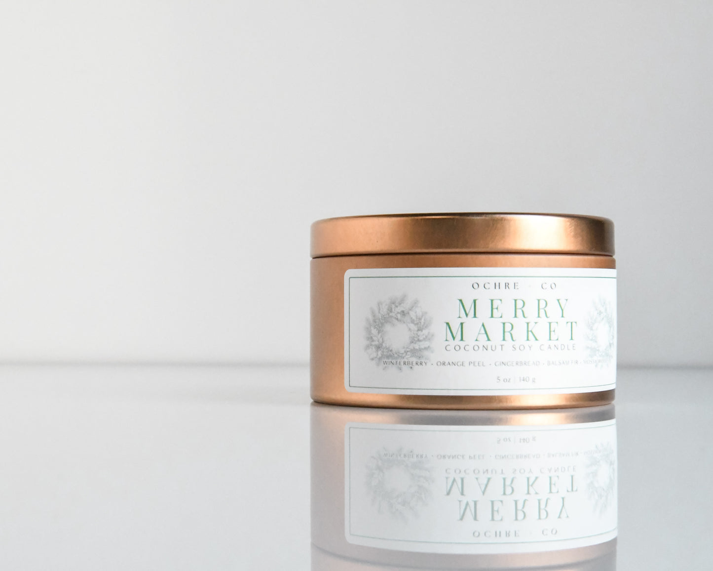 A copper metal candle vessel with the label 'Merry Market' on a white reflective surface against a white background.