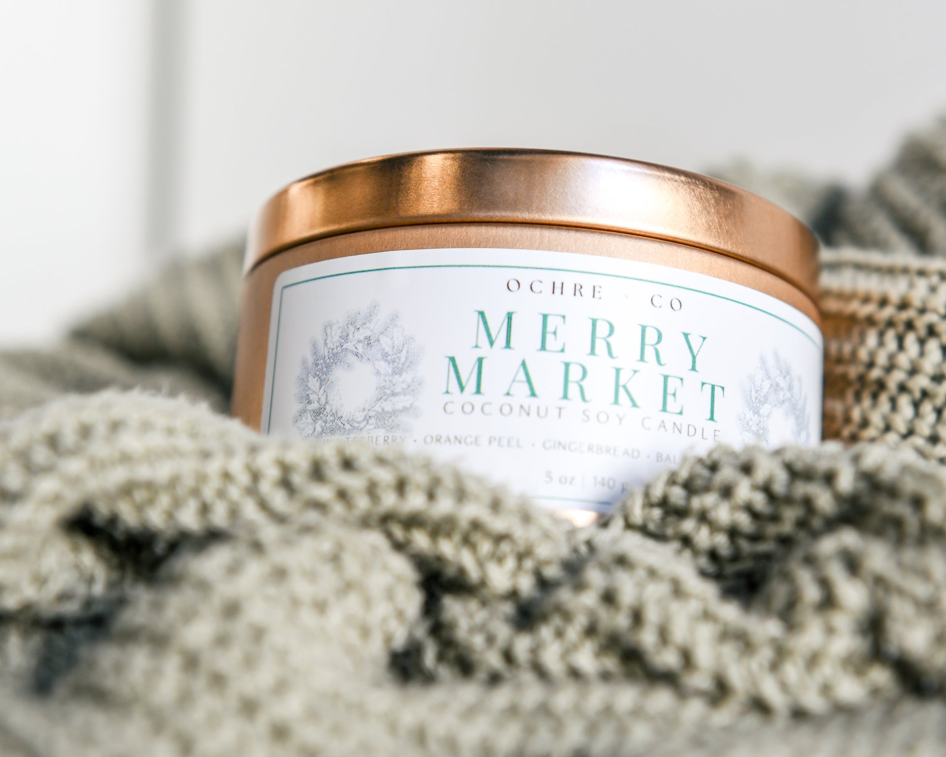 A copper metal candle vessel "Merry Market" rests in a sage-colored sweater with a white background