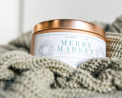 A copper metal candle vessel "Merry Market" rests in a sage-colored sweater with a white background