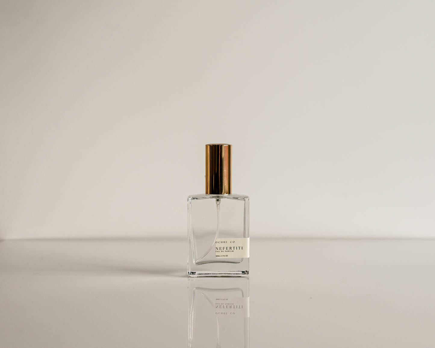 Clear perfume bottle with gold cap on a reflective surface and light gray background