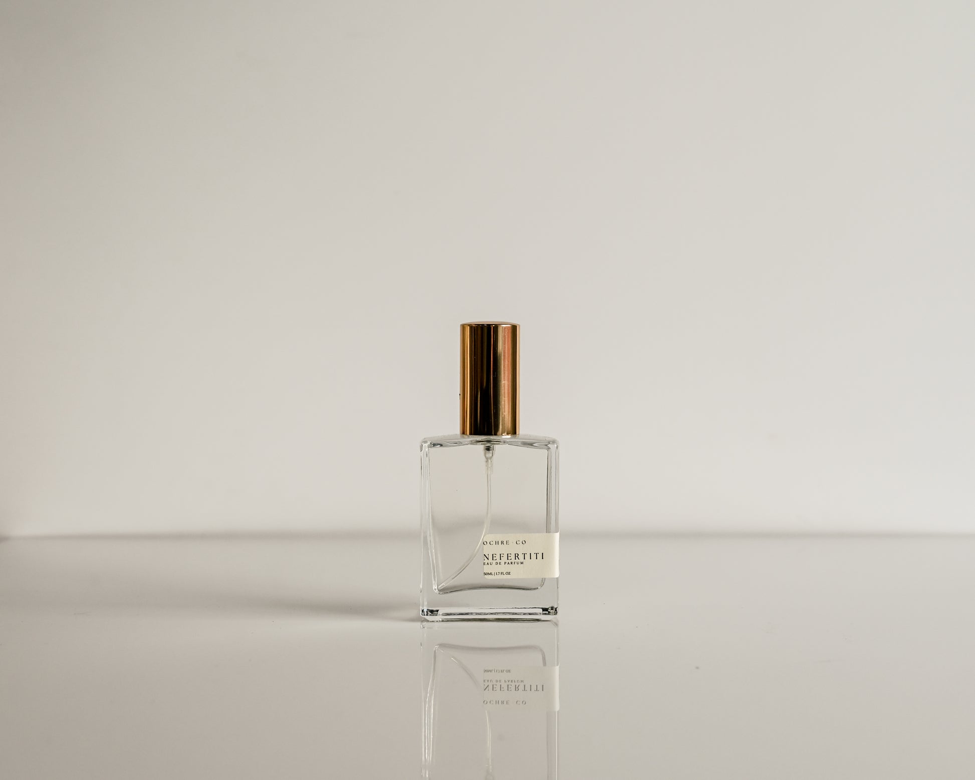 Clear perfume bottle with gold cap on a reflective surface and light gray background