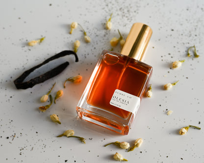 Perfume bottle of Olesia with gold cap and orange liquid on a gray surface with dried flowers and a vanilla bean.