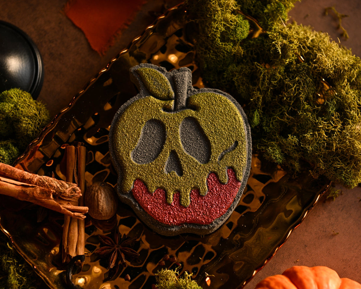A Halloween-themed apple-shaped bath bomb decorated with a dark gray skull face, olive green "poison," and red apple skin showing underneath, displayed on a decorative tray with moss, cinnamon sticks, and a small pumpkin.